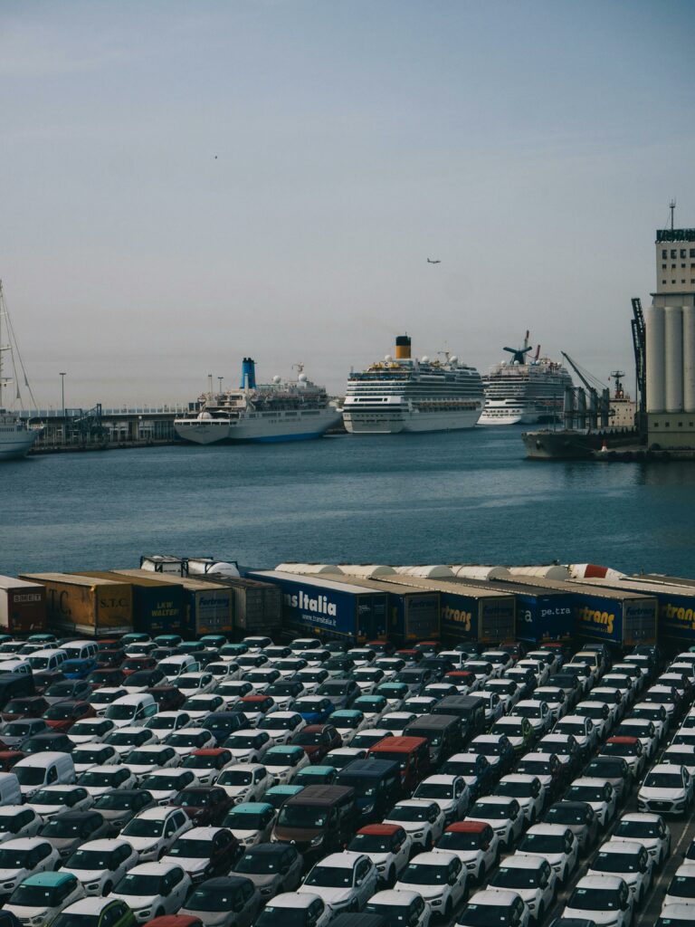 Cargo and cruise ships docked at Barcelona port with rows of parked cars in foreground.